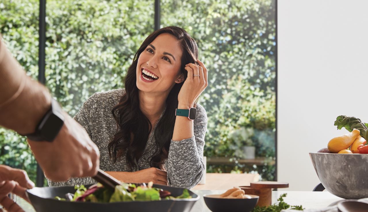 Frau am Tisch sitzend mit Versa 2 Smartwatch Frau am Tisch sitzend mit Versa 2 Smartwatch